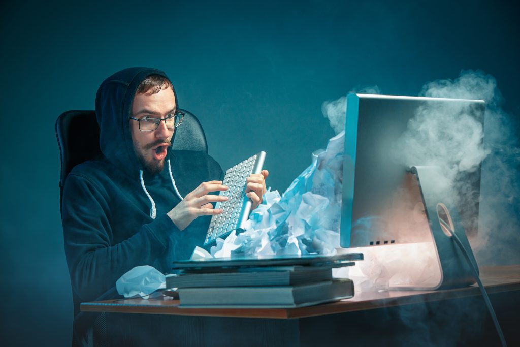 Young stressed handsome businessman working at desk in modern office shouting at laptop screen and being angry about e-mail spam. Collage with a mountain of crumpled paper. Business, internet concept